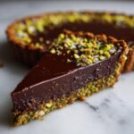 A close-up slice of a rich chocolate pistachio tart showing a glossy ganache filling and a vibrant green pistachio crust.