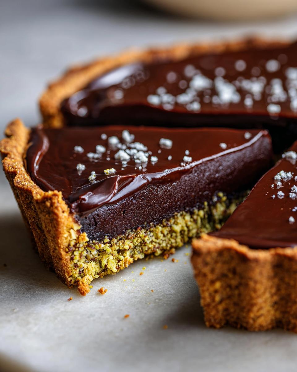 Close-up of a slice of rich chocolate pistachio tart showing the dark ganache and green pistachio crust, topped with flaky sea salt.