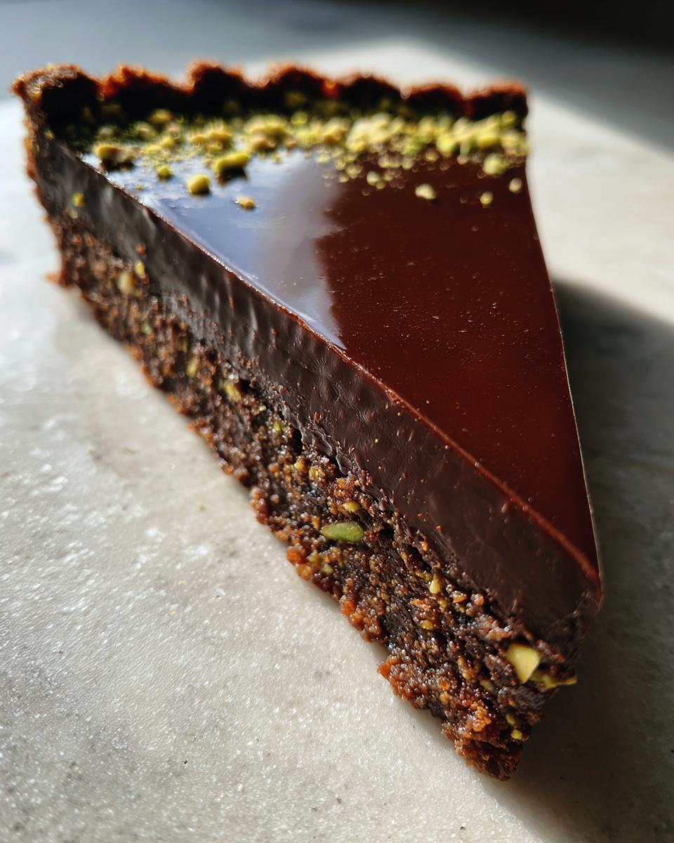 A close-up slice of a rich chocolate pistachio tart showing a dense, nutty crust and a shiny, dark chocolate ganache topping.