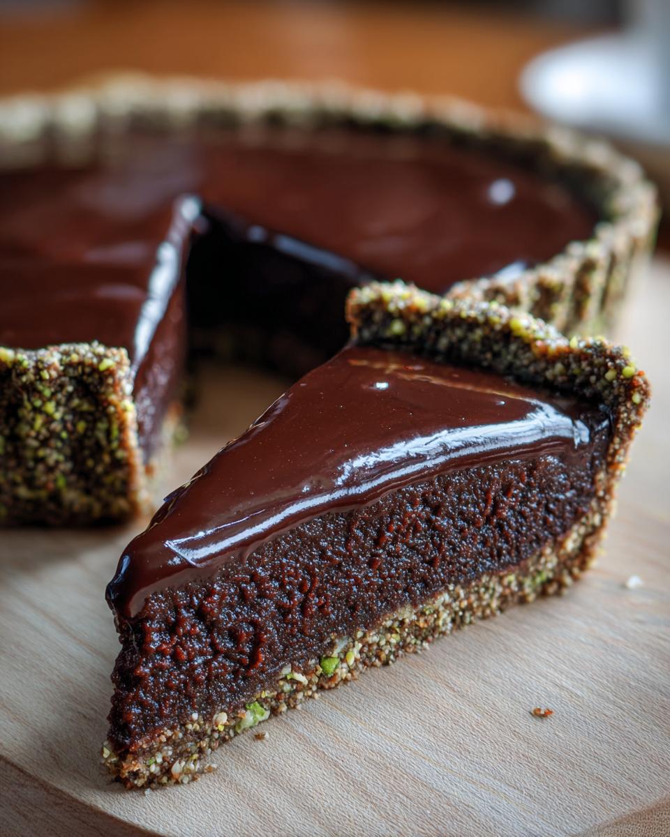 A close-up of a slice of rich chocolate pistachio tart showing the dark filling and shiny ganache topping.