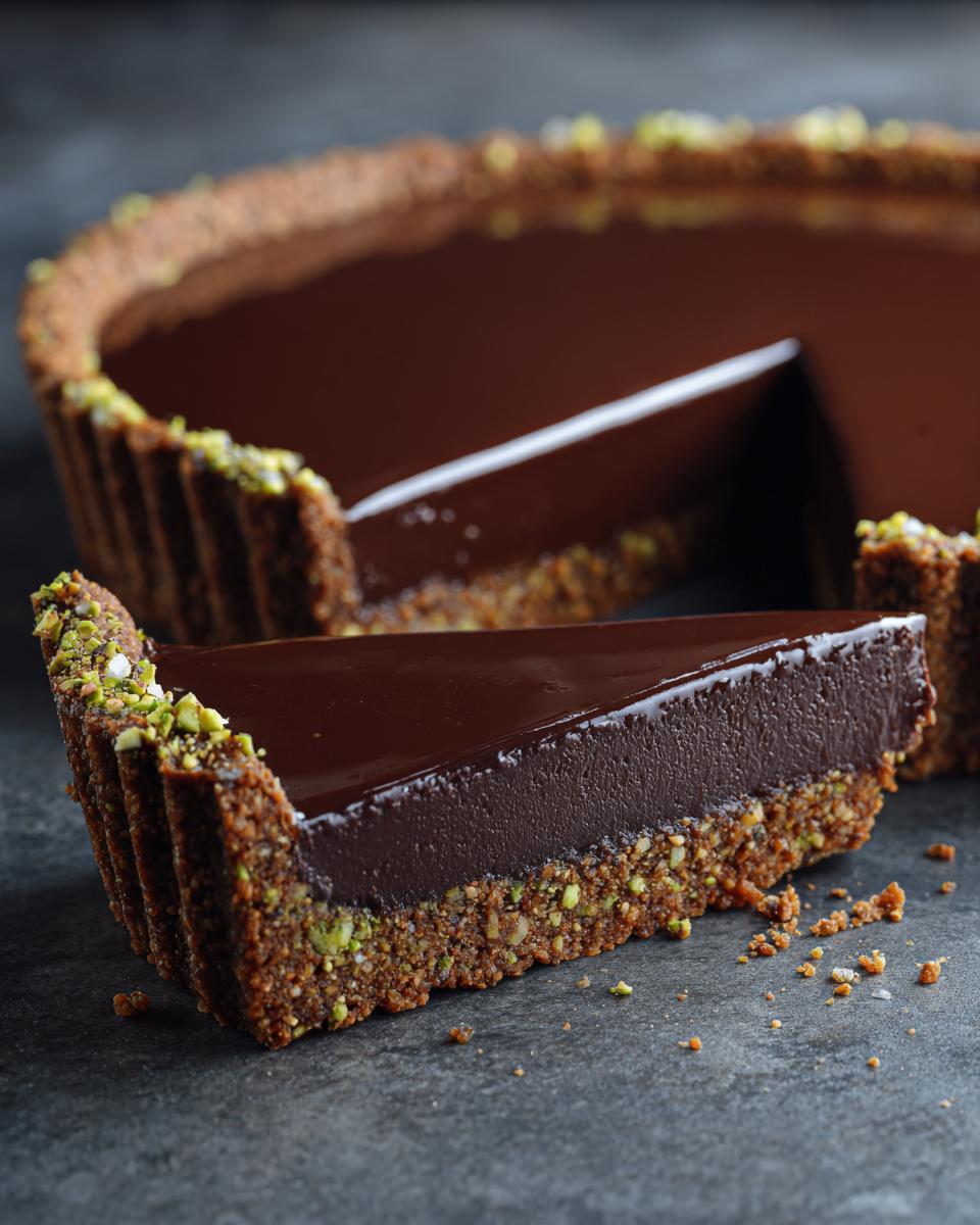 A slice of rich chocolate pistachio tart featuring a thick, glossy chocolate filling and a nutty crust, garnished with chopped pistachios.