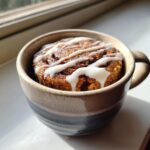 A freshly baked cinnamon mug cake topped with white vanilla icing, sitting in a ceramic mug by a sunny window.