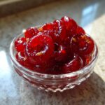 Close-up of glistening, deep red cranberry jalapeño cowboy candy pieces packed tightly in a small, clear glass bowl.