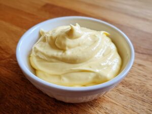 A close-up of thick, creamy homemade mayonnaise recipe eggless, swirled in a small white bowl on a wooden surface.