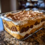 A close-up of a creamy pumpkin trifle dessert showing distinct layers of cake, pumpkin cream, and whipped cream, topped with cinnamon.