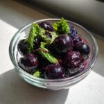 A small glass bowl filled with glossy, dark fermented blueberries with fresh mint leaves on top.