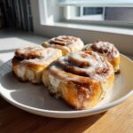 Four freshly baked gluten-free cinnamon rolls generously topped with white icing, sitting on a white plate.