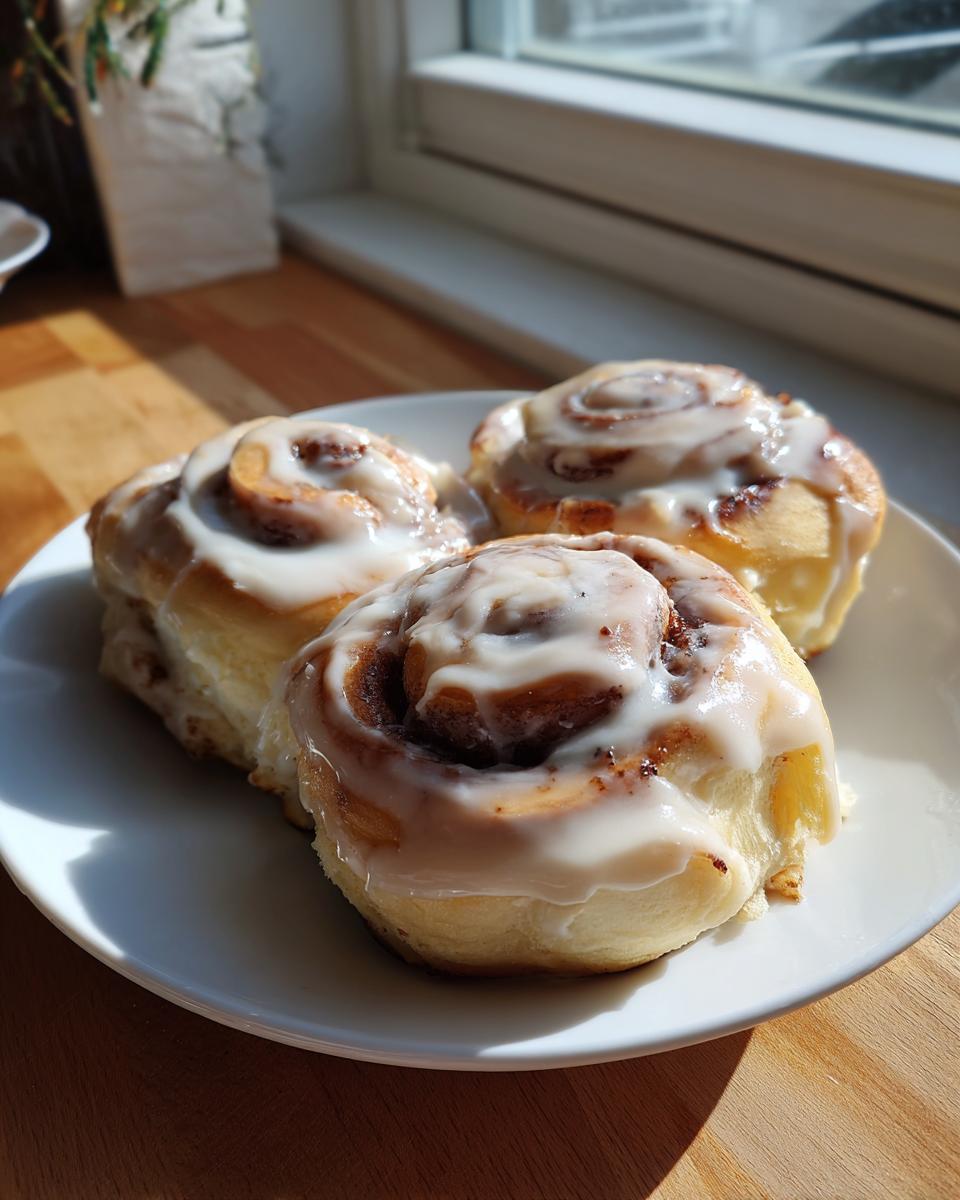 Close-up of three fluffy gluten-free cinnamon rolls covered in white icing, sitting on a white plate near a window.