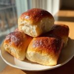A stack of four golden-brown, fluffy sourdough dinner rolls with a shiny glaze, sitting on a white plate.
