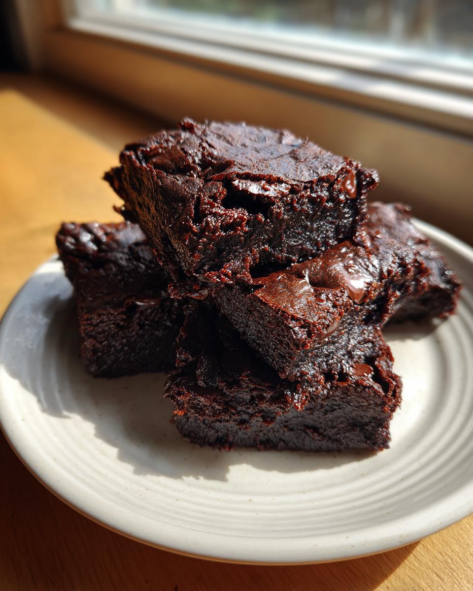 A stack of four rich, fudgy black bean brownies no sugar resting on a light ceramic plate near a window.