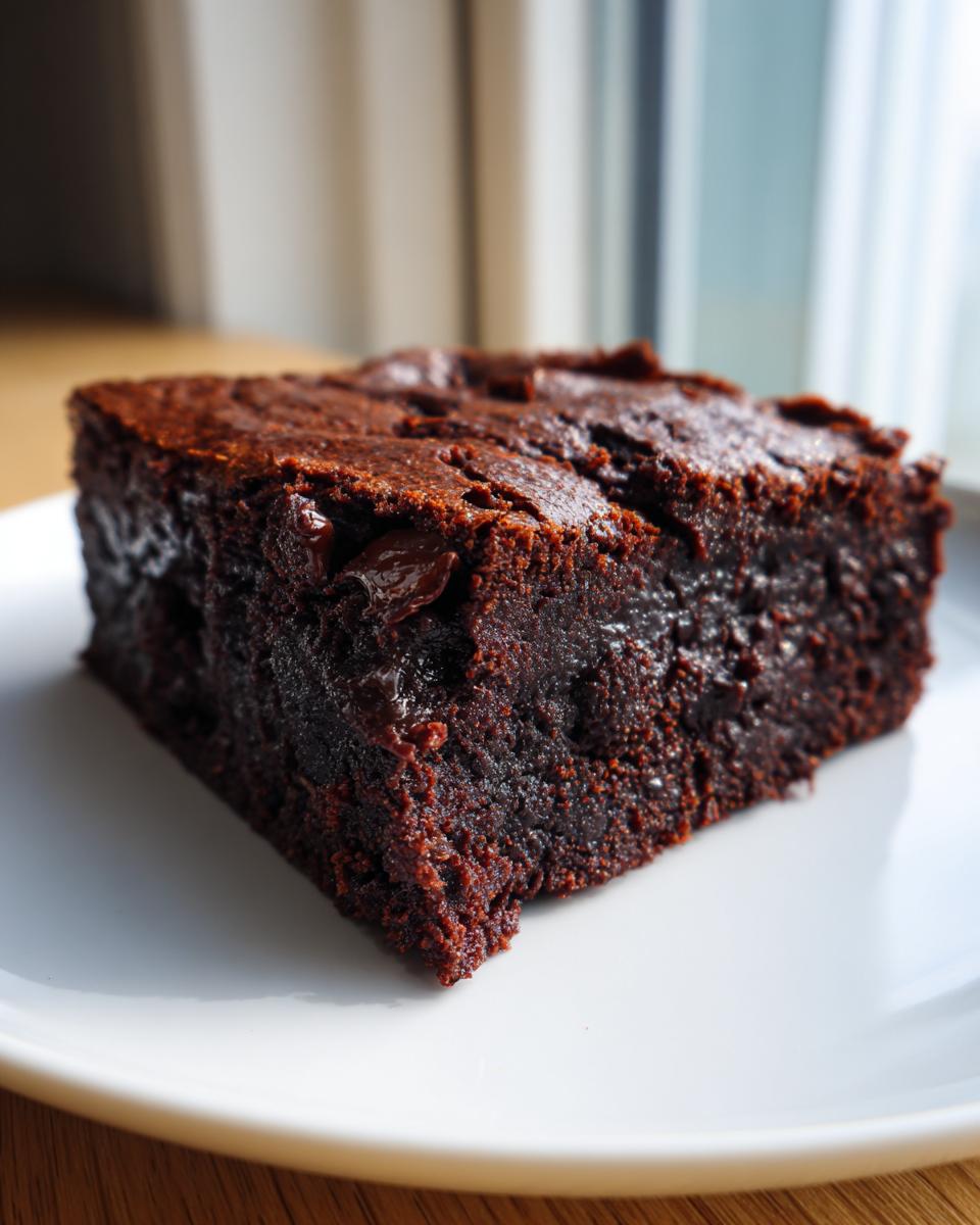 Close-up of a rich, fudgy slice of black bean brownies resting on a white plate near a window.