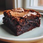 A close-up of a rich, fudgy brownie with a shiny, cracked top sprinkled with flaky sea salt.