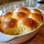 A white baking dish filled with golden brown, glistening fluffy dinner rolls, one roll pulled slightly apart showing the soft interior.