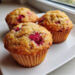 Close-up of three golden baked greek yogurt raspberry muffins topped with coarse sugar on a white plate.