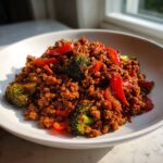 A close-up of a bowl filled with flavorful ground beef stir fry mixed with bright red peppers and green broccoli florets.
