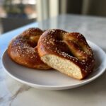 Close-up of two golden-brown high-protein high-fiber pretzels on a white plate, one sliced open showing the soft interior.