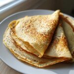 A stack of golden brown, thin high-protein oat & buckwheat crepes folded on a white plate.