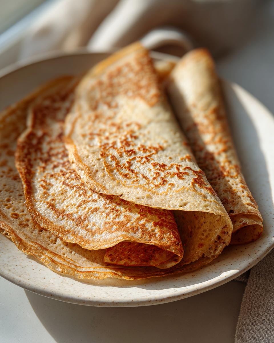 Close-up of golden brown, thin high-protein oat & buckwheat crepes folded and stacked on a speckled white plate.