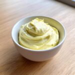 A white bowl filled with thick, creamy, pale yellow homemade mayonnaise recipe avocado oil, sitting on a light wooden surface.