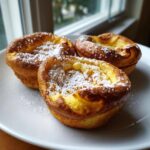 Three golden brown mini dutch baby pancakes dusted generously with powdered sugar, served on a white plate.