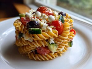 A molded serving of Greek-style pasta salad featuring rotini pasta, cherry tomatoes, cucumber, olives, and feta cheese.