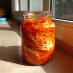 A glass jar filled with vibrant red napa cabbage kimchi, packed tightly and glistening in the sunlight.