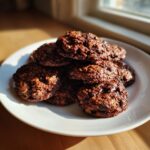 A stack of rich, dark no bake oatmeal peanut butter chocolate chip cookies resting on a white plate near a sunny window.