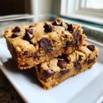 Two thick, chewy peanut butter chocolate chip cookie bars stacked on a white plate, loaded with melted chocolate chips.