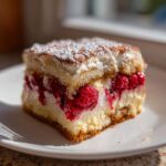 A close-up of a square slice of raspberry tiramisu featuring layers of sponge cake, cream, and fresh raspberries, dusted with powdered sugar.