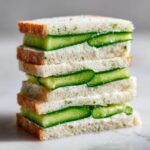 A tall stack of three halved cucumber sandwiches showing layers of white bread, cream cheese spread, and thick cucumber slices.