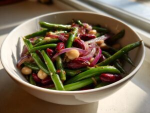 Close-up of a vibrant three bean salad featuring bright green beans, red kidney beans, chickpeas, and sliced red onion in a white bowl.