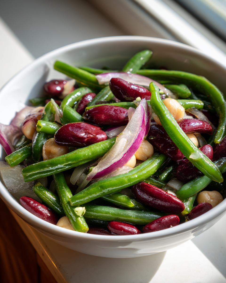 Close-up of a vibrant three bean salad featuring green beans, kidney beans, chickpeas, and red onion slices in a white bowl.