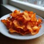 A close-up of a pile of crispy, seasoned air fryer carrot chips served on a light blue plate near a window.