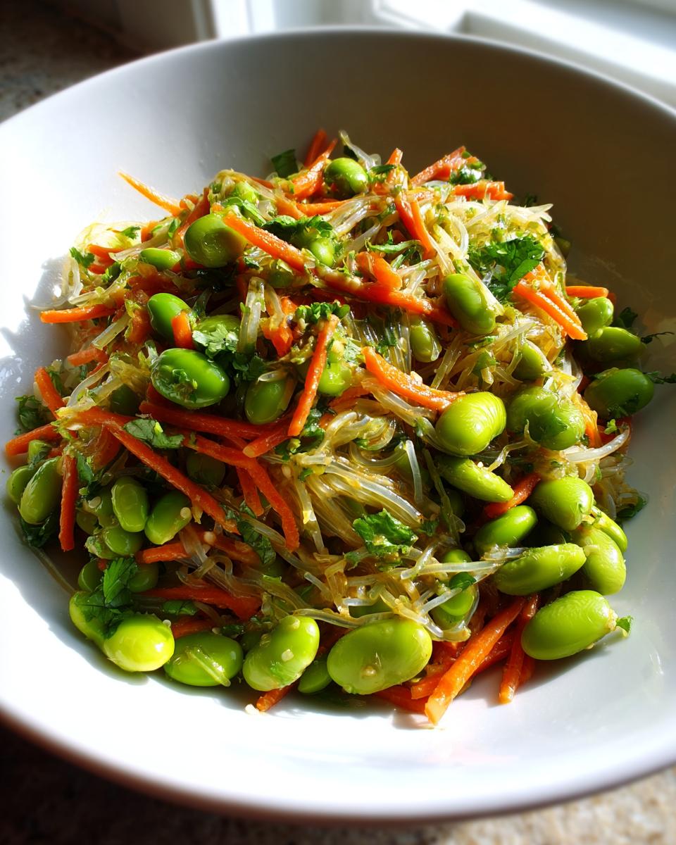 Close-up of a bowl filled with vibrant asian edamame salad featuring bright green edamame, shredded carrots, and clear glass noodles.