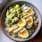 A close-up overhead view of an avocado egg quinoa bowl topped with sliced avocado, soft-boiled eggs, and a creamy white dressing.