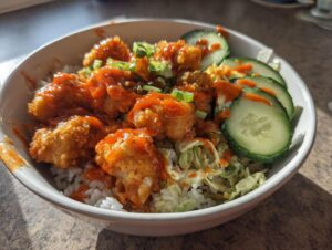 A close-up of a white bowl filled with rice, crispy fried chicken pieces coated in orange sauce, coleslaw, and sliced cucumbers, making up a bang bang chicken bowl.