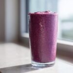 A tall glass filled with a thick, deep purple blueberry yogurt smoothie, showing condensation on the outside.