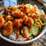 A close-up of a delicious bang bang chicken bowl featuring crispy fried chicken pieces over rice, topped with spicy sauce, cucumbers, and slaw.