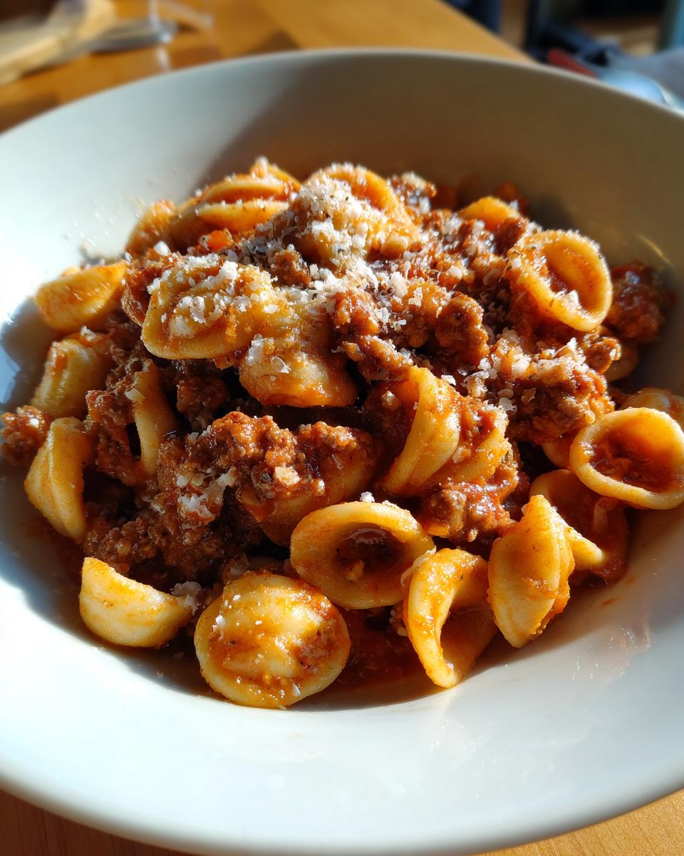 Close-up of a white bowl filled with ground beef orecchiette pasta coated in rich tomato sauce and topped with grated Parmesan cheese.