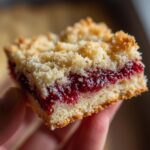 A close-up view of a single, thick raspberry shortbread bars slice held in a hand, showing the crumbly topping and bright red filling.