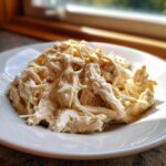 A close-up of creamy, shredded crockpot Olive Garden chicken served on a white plate with natural light from a window in the background.