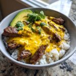 A close-up of a steak queso rice bowl featuring sliced steak, bright yellow queso sauce, white rice, and avocado slices.