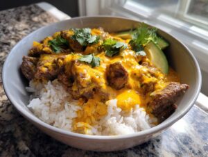 A close-up of a steak queso rice bowl featuring white rice topped with seasoned steak covered in bright yellow queso, garnished with cilantro and avocado slices.