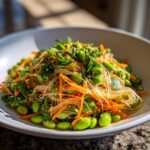 Close-up of a vibrant asian edamame salad featuring glass noodles, shredded carrots, and fresh herbs.