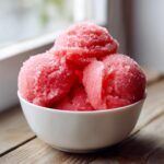 Three scoops of bright pink, icy watermelon sorbet sprinkled with sugar crystals in a white bowl.
